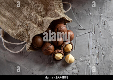 Pile macadamia nuts open kernels and shells in burlap bag on gray concrete background, top view. Stock Photo