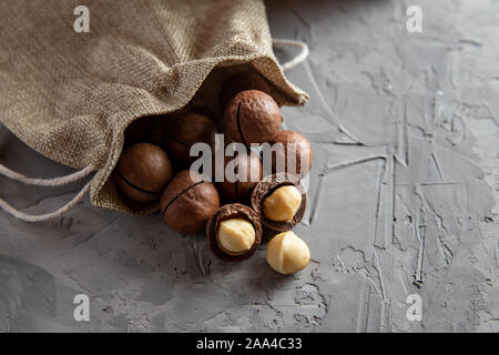 Pile macadamia nuts open kernels and shells in burlap bag on gray concrete background, top view. Stock Photo