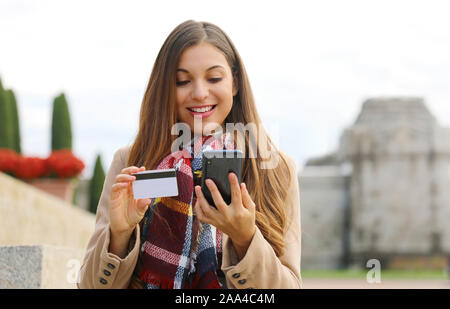 Young shopper woman holding credit card buying online with smart phone sitting outdoor. Stock Photo