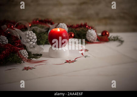 Christmas setting: a red lit candle on foreground surrounded by pine branches, red baubles, red and white ribbons, white pine cones on Christmas table Stock Photo