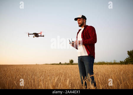 Farmer holds remote controller with his hands while quadcopter is flying on background. Drone hovers behind the agronomist in wheat field Stock Photo