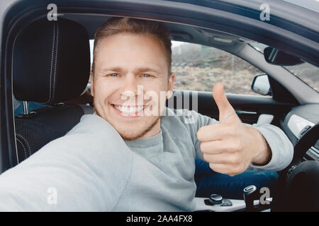 Male driver smiles and takes selfie photo while driving right-hand drive. Car travel rental concept. Stock Photo