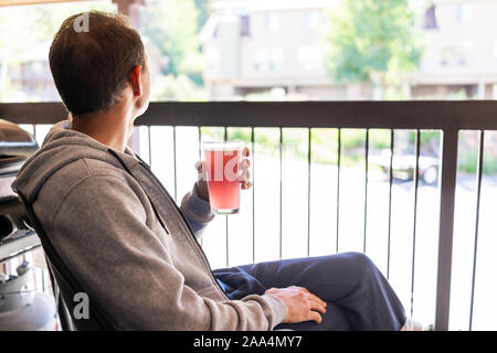 Man sitting outside in chair drinking glass of red pink lemonade cocktail juice on apartment condo balcony in Colorado mountain town looking at view o Stock Photo