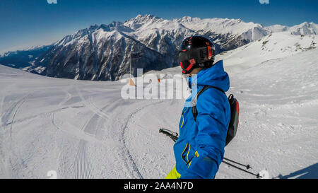 A young skier takes a selfie while skiing down a perfectly groomed slope. Man is wearing helm for the protection. Tall Austrian Alps in the back. Each Stock Photo
