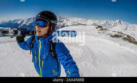 A young skier takes a selfie while carrying his skies up a perfectly groomed slope. Man is wearing helm for the protection. Tall Austrian Alps in the Stock Photo