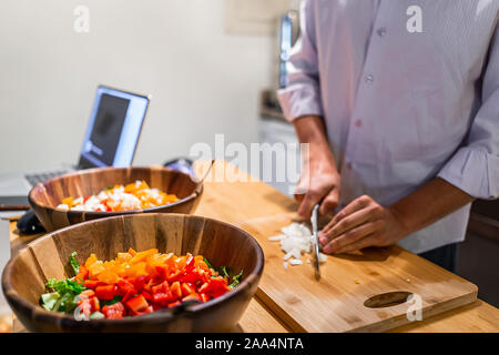 Man chopping onions on cutting board table with two large bowls of fresh vegan salad with red orange bell peppers in kitchen and laptop Stock Photo