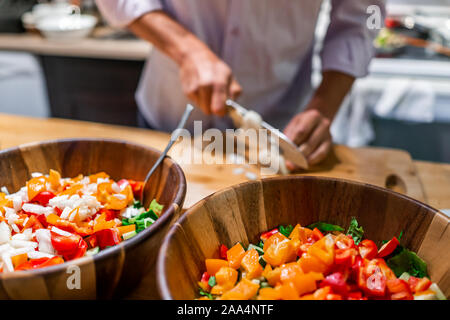 Man chopping onions on cutting board table in background with two large bowls of fresh vegan salad with red orange bell peppers in kitchen Stock Photo