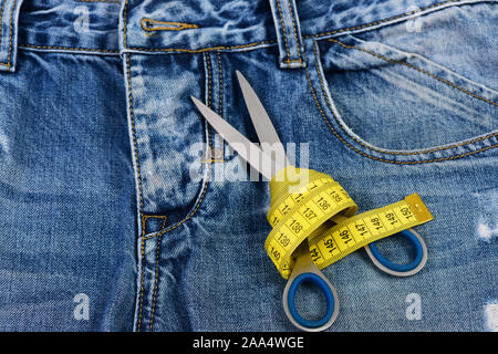 Tailors tools on denim fabric, selective focus. Making clothes and design concept. Jeans crotch, pocket and belt loops, close up. Measure tape wound around metal scissors on jeans. Stock Photo