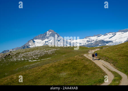 A couple of elderly tourists hiking in Vanoise National Park, French Alps. Beautiful mountain with snowy slopes and clear blue sky on the hohorizon. Stock Photo