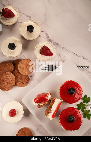 Top view of homemade cheesecakes bathed in strawberry sauce and accompanied with cookies, raspberries, blackberries and leaves on marble background Stock Photo