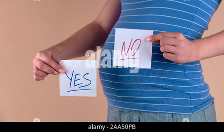 A close-up view of the belly of a pregnant woman in a blue T-shirt, which holds in her hands two sheets with the words YES and NO. The girl chooses NO Stock Photo