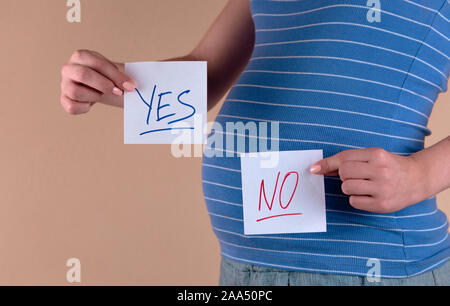 A close-up view of the belly of a pregnant woman in a blue T-shirt, which holds in her hands two sheets with the words YES and NO. The girl chooses YE Stock Photo