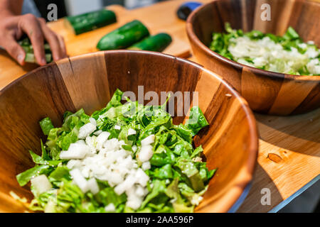 Chopping vegetables on cutting board table with two large bowls of fresh vegan salad with romaine lettuce onions and cucumbers in kitchen Stock Photo