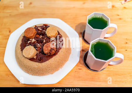 Small yellow vanilla dessert homemade sponge cake with chocolate frosting and macaroon decoration with two cups of matcha green tea on table Stock Photo