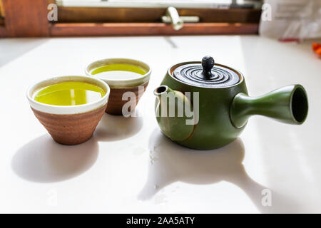 Green clay teapot with kyusu handle on counter by window with Japanese tea brewing genmaicha or sencha in cups during ceremony Stock Photo