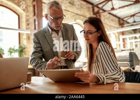 Senior man in formal wear holding coffee cup and explaining something to his young female colleague while working together in the modern office. Busin Stock Photo