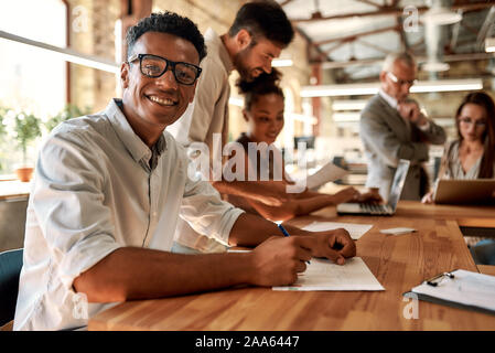 Young and cheerful afro american man in eyeglasses looking at camera with smile while working with his colleagues in the modern office. Business conce Stock Photo