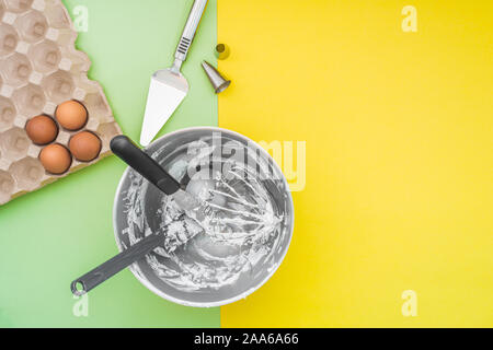 Baking cake utensils in mixer bowl after cake making, nozzles, raw eggs on flat lay green yellow background Stock Photo