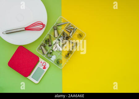 Baking tools accessories on flat lay, green yellow background. Cake turn table, weighing scale machine and a box of piping nozzles. Stock Photo