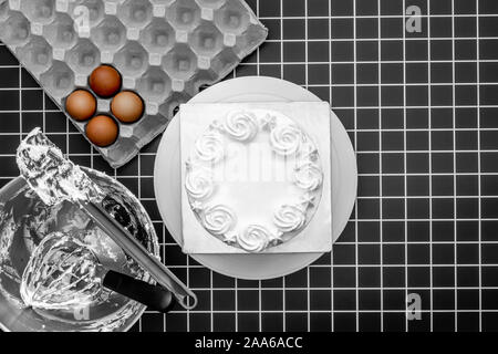 White fresh cream frost cake, baking utensils in mixer bowl, four raw eggs and box of nozzles on flat lay black checked background Stock Photo
