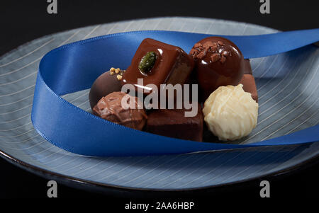 A bunch of exquisite chocolate cubes, embellished with blue ribbons, placed on a dark blue ceramic plate with parallel stripes. Stock Photo