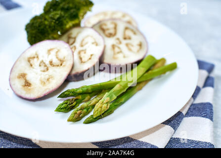 Steamed vegetables in a white plate on a blue table. Eggplant, broccoli, asparagus. Stock Photo