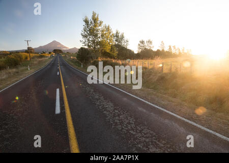 Wide angle view on Mount Taranaki (Mount Egmont) in New Zealand with road leading to it. Taken during sunset with sun rays and lens flare visible. Stock Photo