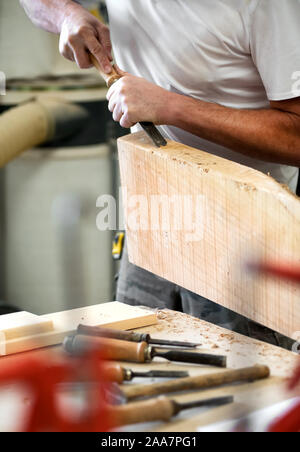 Carpenter working on a block of wood with a chisel in a close up on his hands with assorted hand tools on the workbench in the foreground Stock Photo