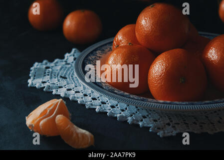 Tangerines on a crochet mat on a black wooden table in a rustic kitchen. Ripe fruit in a dark food photography with empty copy space. Stock Photo