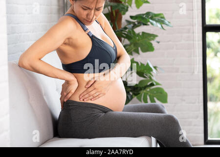 Side view of upset Asian pregnant woman touching back and side suffering from backache and hip pain in light apartment Stock Photo