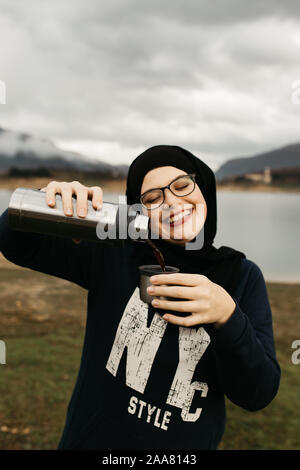 Happy woman with hijab drinking coffee in nature, on a foggy morning Lake and mountains are in the background. She is smiling and poring coffee. Stock Photo