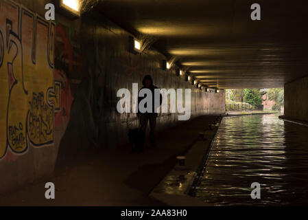 An isolated, hooded, solitary figure walking by an urban canal in dark, UK pedestrian underpass, passing urban graffiti artwork sprayed on walls Stock Photo