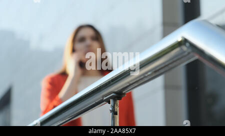 Defocused shoot of a business woman in a red jacket talking on the phone. Selective focus metal fence near the business center. Stock Photo
