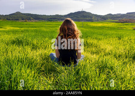 Young caucasian woman red-haired with freckles, resting on a green field at spring sunset, sitting in a yoga position. Meditation, mindfulness, rela Stock Photo