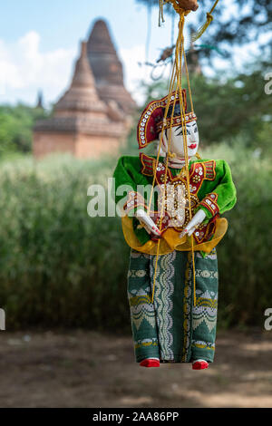 Traditional Burmese wooden puppet dressed in an elaborate costume hangs from a tree with an ancient temple of Bagan, Myanmar in the background Stock Photo