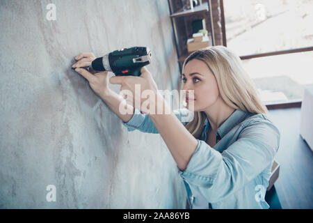 Profile side view portrait of her she nice attractive pretty lovely focused wavy-haired girl making hole in grey wall with drill at modern industrial Stock Photo