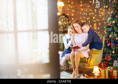 Happy mother reads fairy tales to children while sitting on a sofa near a festive Christmas tree Stock Photo