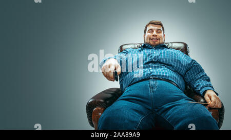 Portrait of fat caucasian man wearing jeanse and whirt sitting in a brown armchair isolated on gradient grey background. Emotional watching TV and changing channels, laughting. Overweight, carefree. Stock Photo