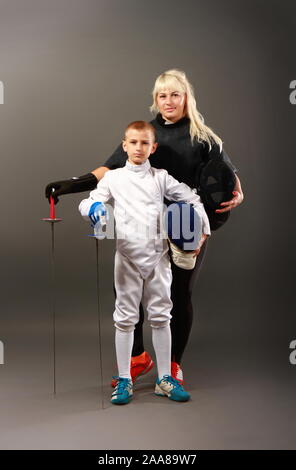 Young blonde girl in a dark coaching outfit and a little boy in a white sports outfit practicing rapier fencing on a gray background Stock Photo