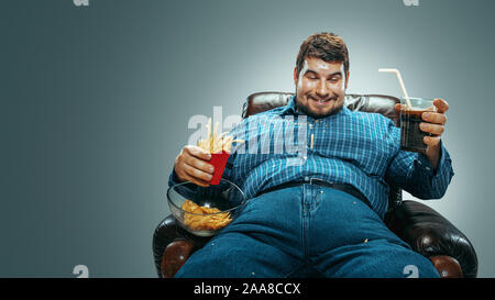 Portrait of fat caucasian man wearing jeanse and whirt sitting in a brown armchair on gradient grey background. Watching TV drinks cola, eats chips, fried potato, laughting. Overweight, carefree. Stock Photo