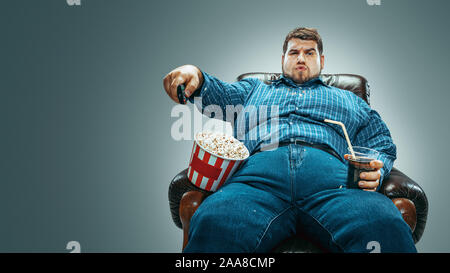 Portrait of fat caucasian man wearing jeanse and whirt sitting in a brown armchair on gradient grey background. Watching TV drinks cola, eats popcorn and changing channels. Overweight, carefree. Stock Photo