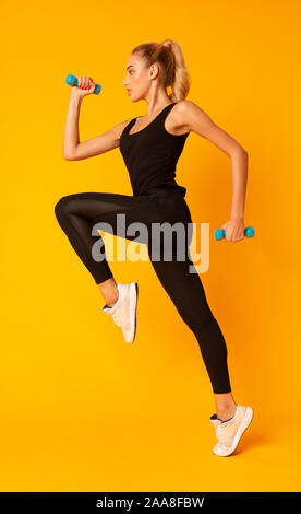 Fitness Girl Jumping Holding Dumbbells Over Yellow Background, Studio Shot Stock Photo