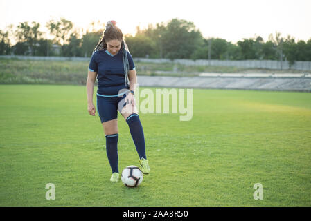 young girl with dreadlocks. In soccer uniform, holds the ball. Overweight woman, an alternative to sports. Losing weight. On the football field, green Stock Photo