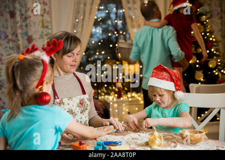 Mother and kids in red hats cooking gingerbread cookies and decorating glaze. Father with child decorating Christmas tree. Beautiful living room Stock Photo