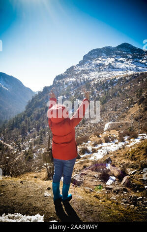 A woman in winter clothing standing on top of the rock of a snowcapped rocky mountain. Rear view. Deep Snow and Blizzard all around. Human face to fac Stock Photo