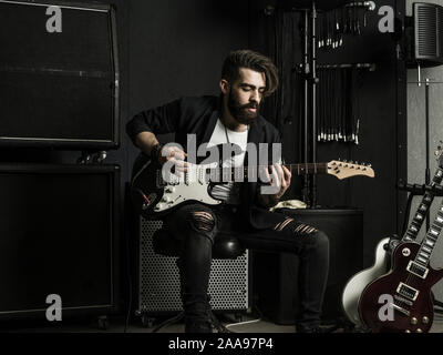 Photo of a man with beard sitting and playing his electric guitar in a recording studio. Stock Photo