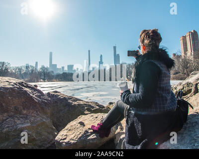Young woman sitting on rocks by frozen lake in Central Park taking pictures of Manhattan Stock Photo