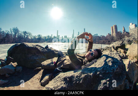 Young woman making heart shape with hands lying on rocks by frozen lake in Central Park with a view of Manhattan Stock Photo