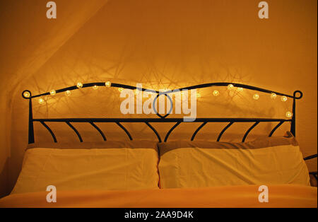 Bedroom interior with comfortable bed and pillows, garland of Christmas lights around the wrought iron headboard. Valentine’s day date concept. Stock Photo