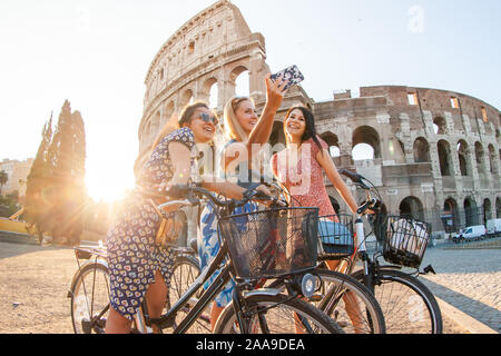 Three happy young women friends tourists with bikes taking selfies at Colosseum in Rome, Italy at sunrise. Stock Photo